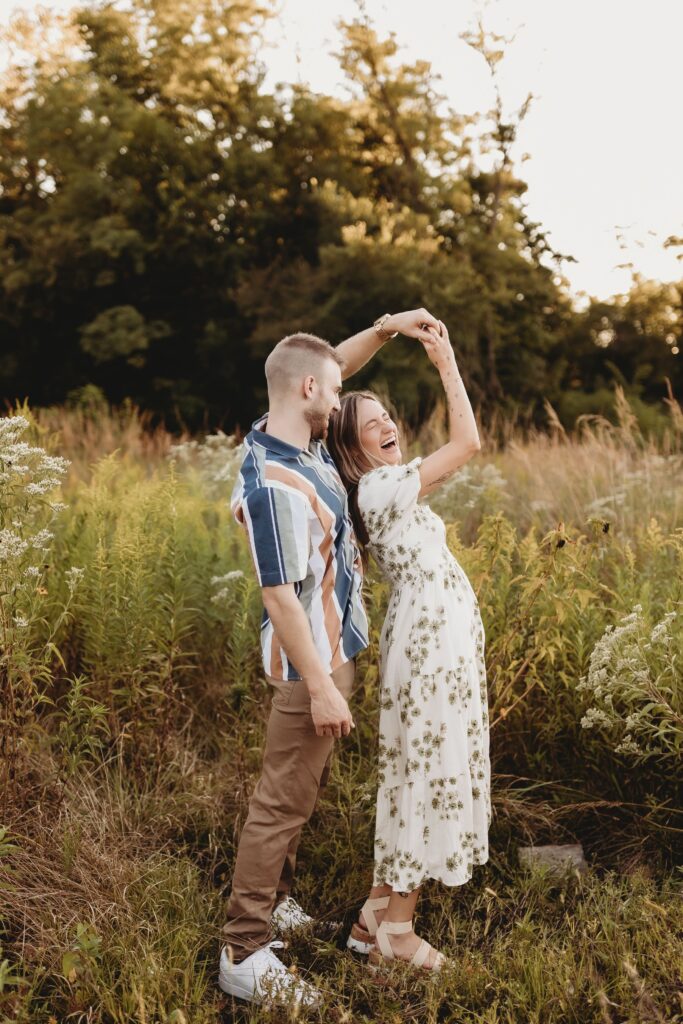 A joyful couple dancing in a sunlit meadow surrounded by tall grass and wildflowers. The woman is wearing a floral dress and the man is dressed in casual summer attire.