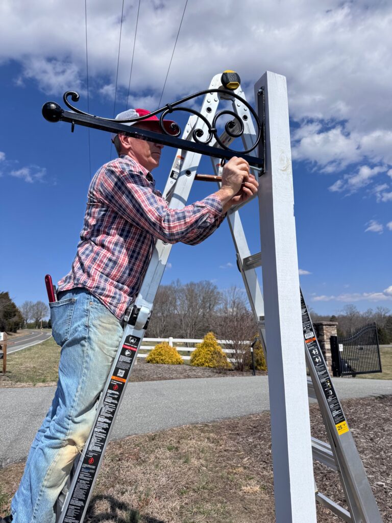 An owner of Willow and Stone Events installs a decorative entry sign bracket under a bright blue sky, enhancing the venue’s charm.