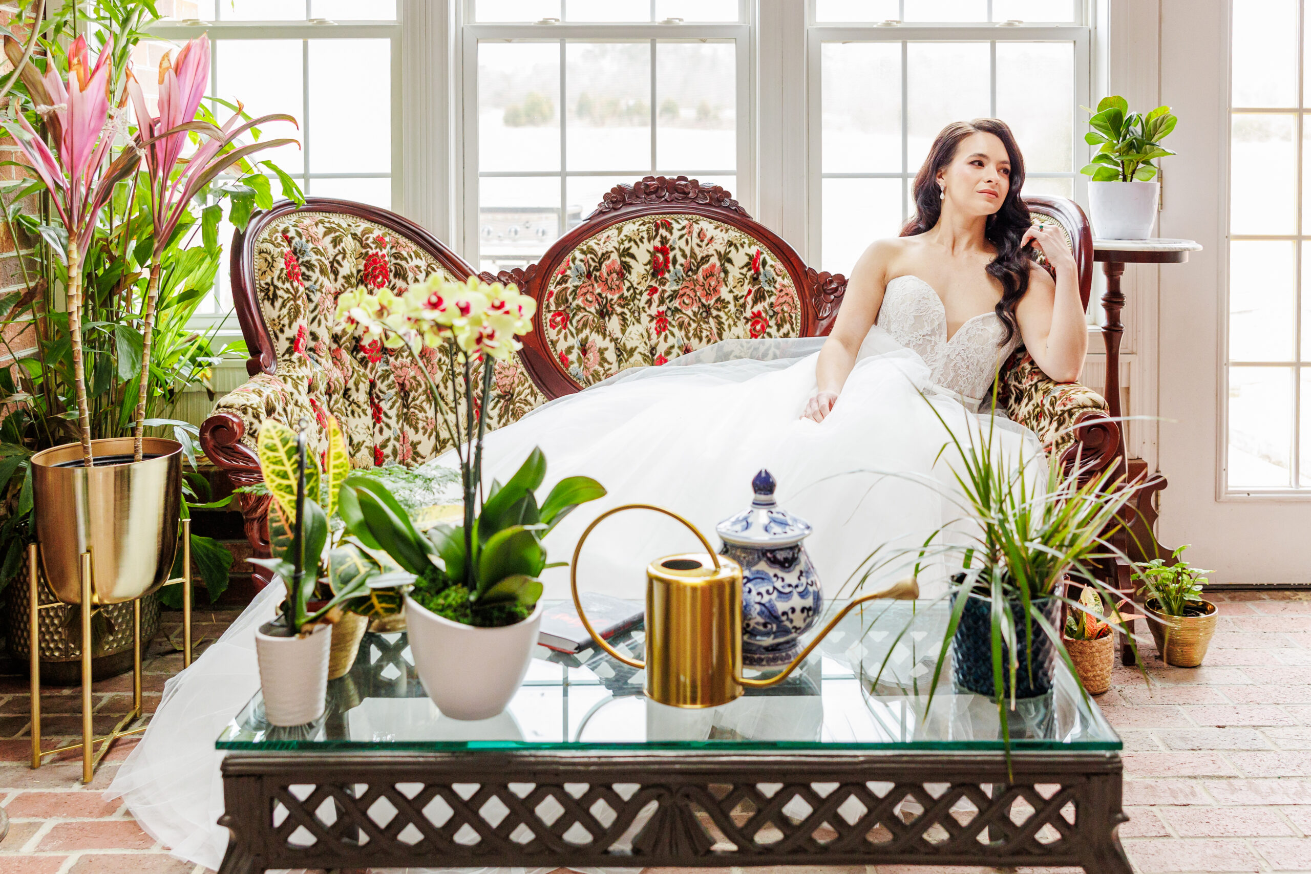 Bride lounging in vintage floral loveseat surrounded by elegant plants in the sunlit solarium at Willow and Stone Events